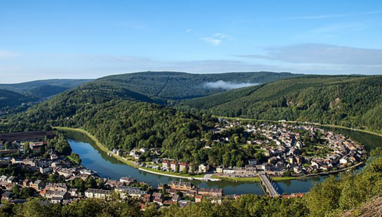Vue du haut d'un colline de la rivière de la Meuse et d'une petite ville, en contrebas, encerclée de collines boisées.